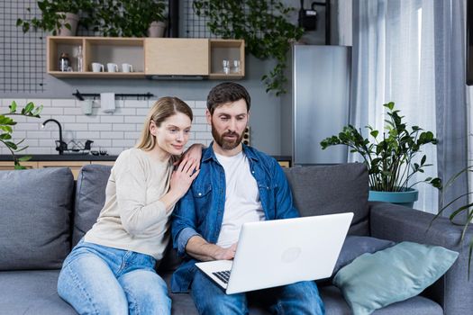 Happy young married couple man and woman together at home, sitting on sofa smiling and using laptop