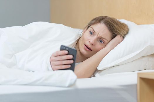 Woman lying in bed in the morning, shocked reading bad news from the phone