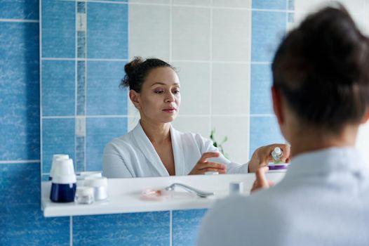 Charming woman in white bathrobe, reflecting in mirror, squeezing some moisturizer during daily procedures in bathroom
