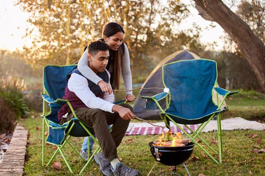 Where theres smoke theres dinner. a young couple roasting marshmallows while camping outside.