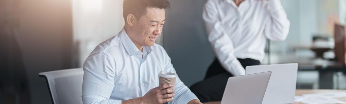 Asian businessman drinking coffee and working on laptop with colleague in background