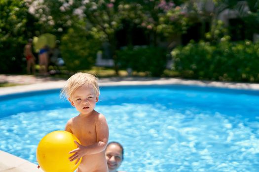 Small baby with a yellow ball stands by the pool with turquoise water