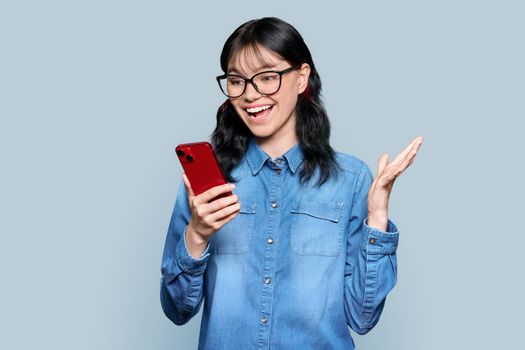 Surprised teenage girl looking at smartphone screen on gray color studio background. Attractive emotional young female student in glasses holding phone in hands. Emotions, mobile app, lifestyle, youth