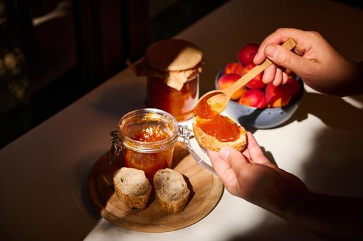 Daylight falls on the table. Hands using wooden spoon, spreads a homemade confiture on the bread. Rustic still life