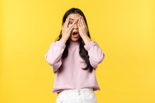 People emotions, lifestyle and fashion concept. Scared and shocked, terrified asian woman screaming in horror, hold hands on face and peeking through fingers, yellow background