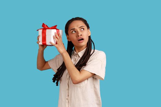Woman with black dreadlocks shaking wrapped present box, being interested what inside, looking away.