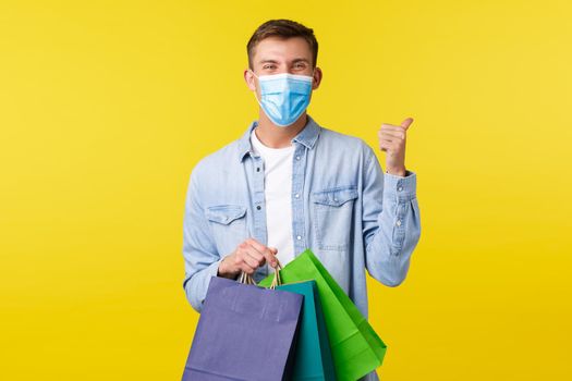 Concept of covid-19 pandemic outbreak, shopping and lifestyle during coronavirus. Happy handsome blond man in medical mask, rejoicing over opened malls, show thumbs-up and carry bags.
