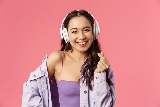 Close-up portrait of attractive happy young modern asian girl, listening music in headphones, show korean heart sign, enjoying favorite music or podcast, bought earphones with discount