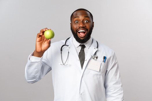 Healthcare, medicine and healthy lifestyle concept. Enthusiastic, amused happy african-american doctor advice eat fruits, showing apple and smiling, persuade kid eat vitamins, grey background