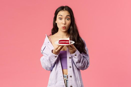 Portrait of excited and surprised cute asian girlfriend holding piece of cake, order delivery desserts from favourite cafe, holding it on plate, fold lips say wow, standing amazed pink background