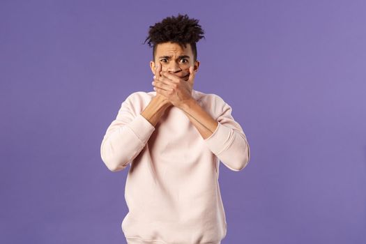 Shocked worried, embarrassed young man said something he shouldnt have, shut his mouth with hands and look guilty or anxious at camera, feel sorry for being rude, purple background
