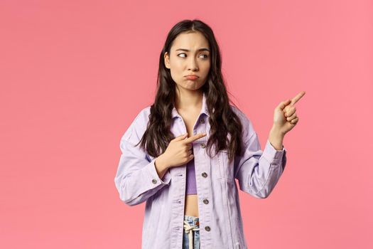 Attractive, gloomy mixed-race girl pointing fingers right, looking at something with regret and sigh, pouting displeased, jealous of missing good opportunity, want be there, pink background