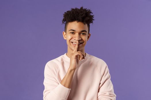 Close-up portrait of attractive smiling young man hiding secret, asking keep silent or quiet to do surprise, show shush gesture, place index finger to mouth, standing purple background
