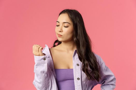 Close-up portrait of arrogant stylish young asian girl not listening, feeling careless and uninterested in conversation, bored looking at hand, thinking visit nails studio, pink background