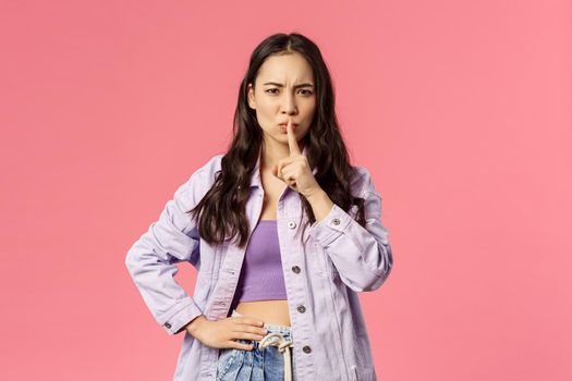 Shush dont speak. Serious-looking asian girl hush, frowning and looking upset, irritated with person speaking too loud, press index finger to lips, scolding, tell keep secret, pink background