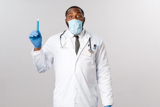 Covid-19, coronavirus patient treatment and disease concept. Excited african-american male doctor in face mask and latex gloves, hold vial with vaccine, invented cure in laboratory