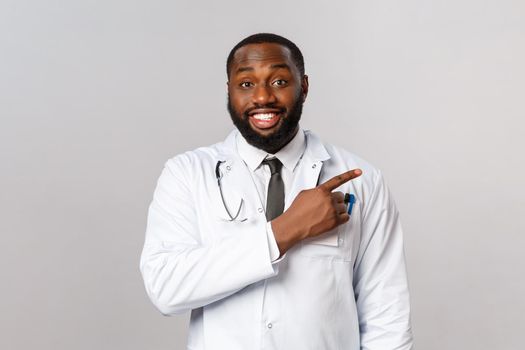 Healthcare, medicine and epidemic concept. Cheerful, friendly-looking african-american male physician explain patients prevention of getting covid19 symptoms, poiting finger right, recommend product