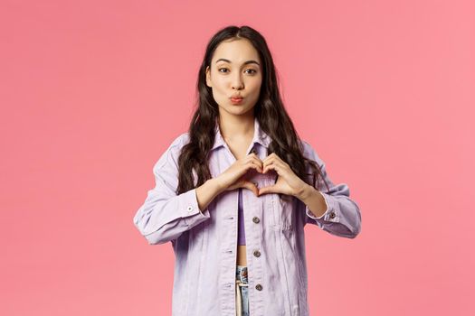 Portrait of romantic cute korean girl show heart gesture and fold lips, giving kiss, express sympathy, passion and romance, standing pink background in lovely stylish outfit