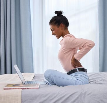Be aware of your posture. Full length shot of an attractive young woman holding her back in pain while working on her laptop in the bedroom at home.
