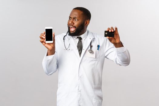 Covid19, pandemic and online medicine concept. Complaining angry african-american man looking at mobile phone display, hold credit card, outraged with huge bills and expensive app subscribtion