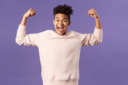 Portrait of young happy man got scholarship, applied to cool university, raise hands up flex biceps like champion, triumphing from great news, achieve goal and rejoicing, purple background