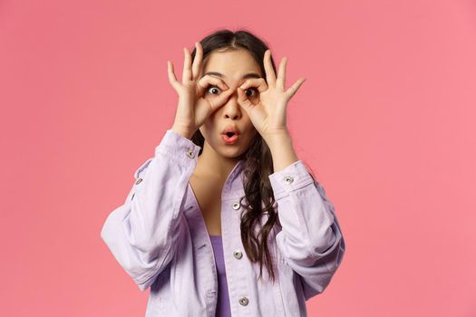 Close-up portrait of amused, speechless and impressed asian young girl seeing something curious and amazing, make glasses with fingers over eyes, fold lips say wow astonished, pink background