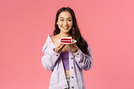 Got you some sweet dessert. Portrait of cheerful young asian girl holding piece of delicious cake, smiling happy, eating out at favorite cafe, standing pink background joyful