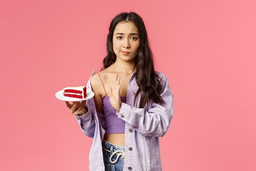 Portrait of skeptical young asian stylish slim girl refusing eat cake, smirk show top sign to dessert lying on plate, rejecting try it, disappointed with bad taste, stand pink background