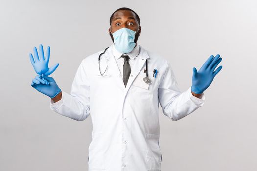 Covid-19, coronavirus patient treatment and disease concept. Funny african-american male doctor in facial mask, protect from virus outbreak, playing with small kid, blowing latex glove