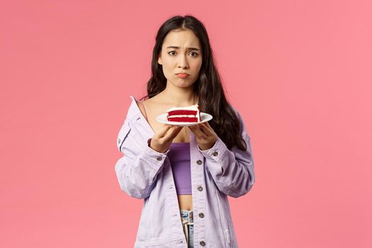 Portrait of gloomy sad cute asian girl just being dumped trying eat-out her sadness, holding piece cake, pouting and frowning upset, have uneasy feelings, grieving, pink background