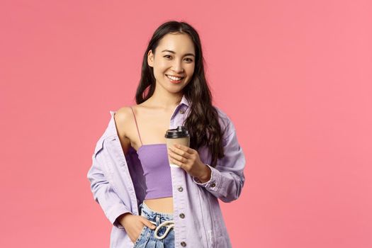Lifestyle, modern people and beauty concept. Sassy good-looking asian girl chilling with friends at favorite cafe, grab take-away beverage, drinking coffee and smiling camera, pink background