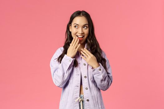 Lifestyle, spring and beauty concept. Portrait of stylish young pretty girl have an idea, giggle, smile and cover mouth, look upper left corner devious and thoughtful, have plan, pink background