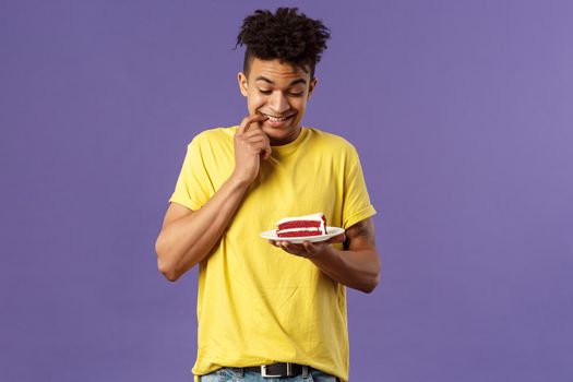 Celebration, party and holidays concept. Portrait of boyfriend cant resist temptation to eat last piece of cake, biting lips and smiling eager to have bite of dessert, hesitating, purple background