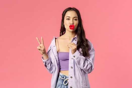 Holidays, lifestyle and people concept. Portrait of sassy good-looking, feminine asian woman in stylish outfit, holding big lips stick and show peace sign, standing pink background