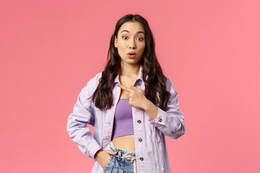 Portrait of surprised and curious young stylish female student pointing finger left and look amazed camera, asking question about interesting thing located there, pink background
