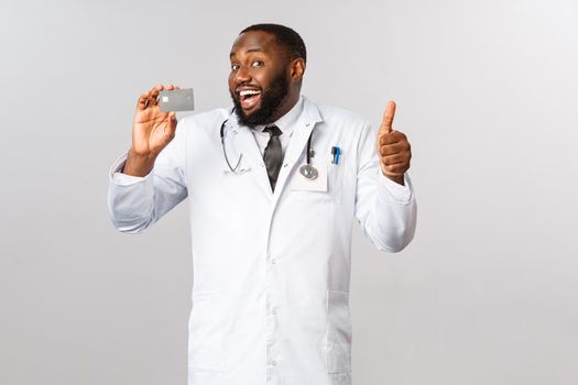 Good-looking pleased male doctor, physician in white coat, holding credit card and show thumb-up in approval or like, recommend bank, online purchases or contactless payment for hygiene purpose