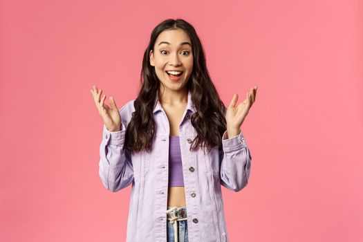 Portrait of surprised, enthusiastic korean girl in denim jacket, look wondered and happy, hear great news, applause, raise hands to clap and praise great work, smiling joyfully, pink background