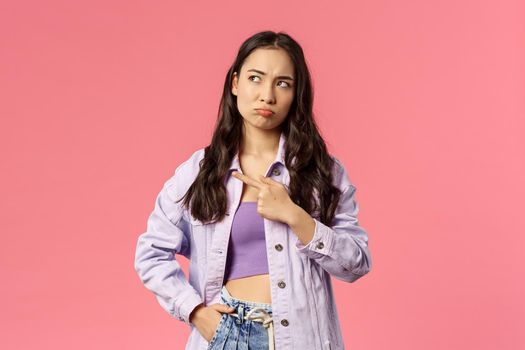 Portrait of complicated cute young korean girl in stylish outfit, frowning looking and pointing upper left corner, standing thoughtful, facing problem thinking of solution, pink background