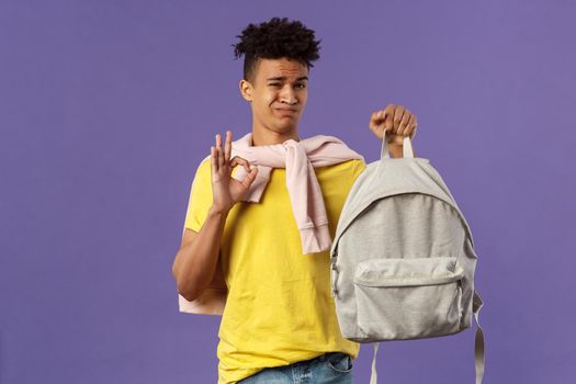 Good but not the best. Portrait of disappointed, unsatisfied young teenage student, guy grimacing without enthusiasm show okay, received new ugly backpack, standing purple background