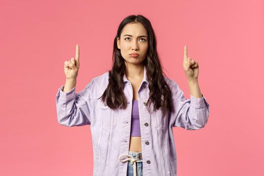 Portrait of gloomy and displeased, young frowning girl in denim jacket, grimacing looking and pointing fingers up disappointed, express regret or jealousy, standing pink background