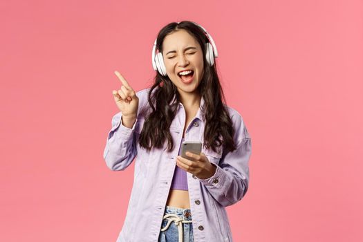 Waist-up portrait of excited and carefree stylish woman enjoying favourite songs, listening music in headphones, close eyes dancing to rhythm and singing along, hold smartphone
