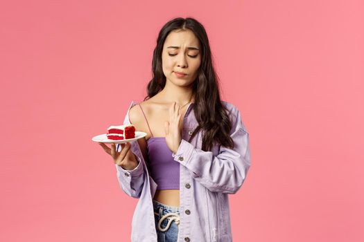 No thank you calories. Portrait of attractive asian girl taking care of her looks, saying no to delicious dessert, smirk show stop sign to cake, restrain herself eat after six, pink background