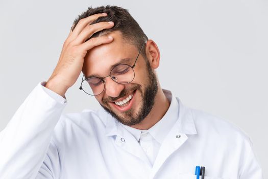 Healthcare workers, coronavirus, covid-19 pandemic and insurance concept. Headshot of handsome young doctor in white coat and glasses, touching hair and laughing, smiling pleased