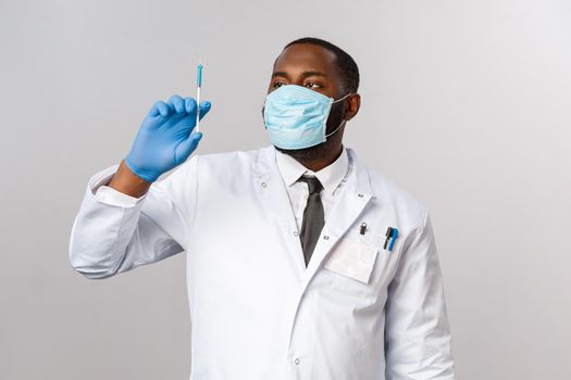 Covid19, pandemic and healthcare concept. Serious handsome african-american doctor in face mask, latex mask, looking at syringe with virus vaccine, treating patients, grey background