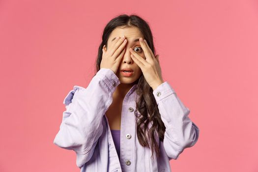 Close-up portrait of scared and alarmed young asian woman panicking, cover eyes from fear, peeking with one eye at something shocking and embarrassing, stand pink background