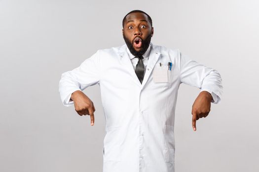 Healthcare, insurance and clinic concept. Amazed, excited african-american doctor telling to hurry up and look down, pointing fingers at bottom, medical product, breaking news