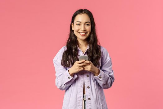 Online lifestyle, people and beauty concept. Cheerful stylish young girl using mobile phone and smiling pleased at camera, chatting during lockdown, use education app, pink background