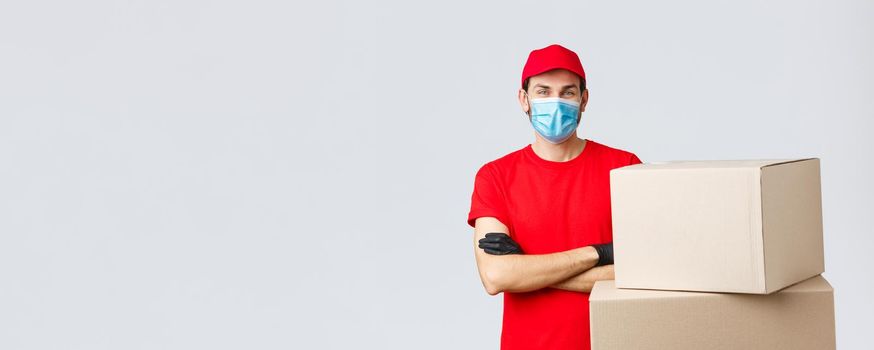 Packages and parcels delivery, covid-19 quarantine and transfer orders. Confident young courier in red uniform, gloves and medical mask, cross arms as standing boxes, grey background