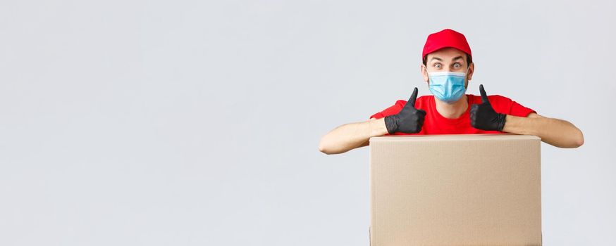Packages and parcels delivery, covid-19 quarantine and transfer orders. Enthusiastic courier in red uniform, face mask and gloves, lean on box order and thumb-up, recommend service, express shipping
