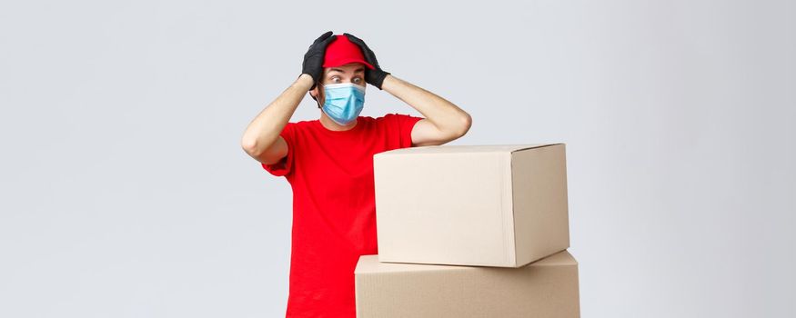 Packages and parcels delivery, covid-19 quarantine and transfer orders. Concerned and troubled courier in red uniform, face mask and gloves, grab head and gasping shocked staring at boxes